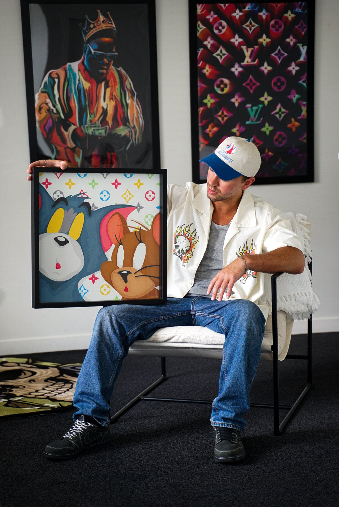 Man sitting on a chair holding a framed artwork with colorful design, surrounded by other artworks on the wall.

